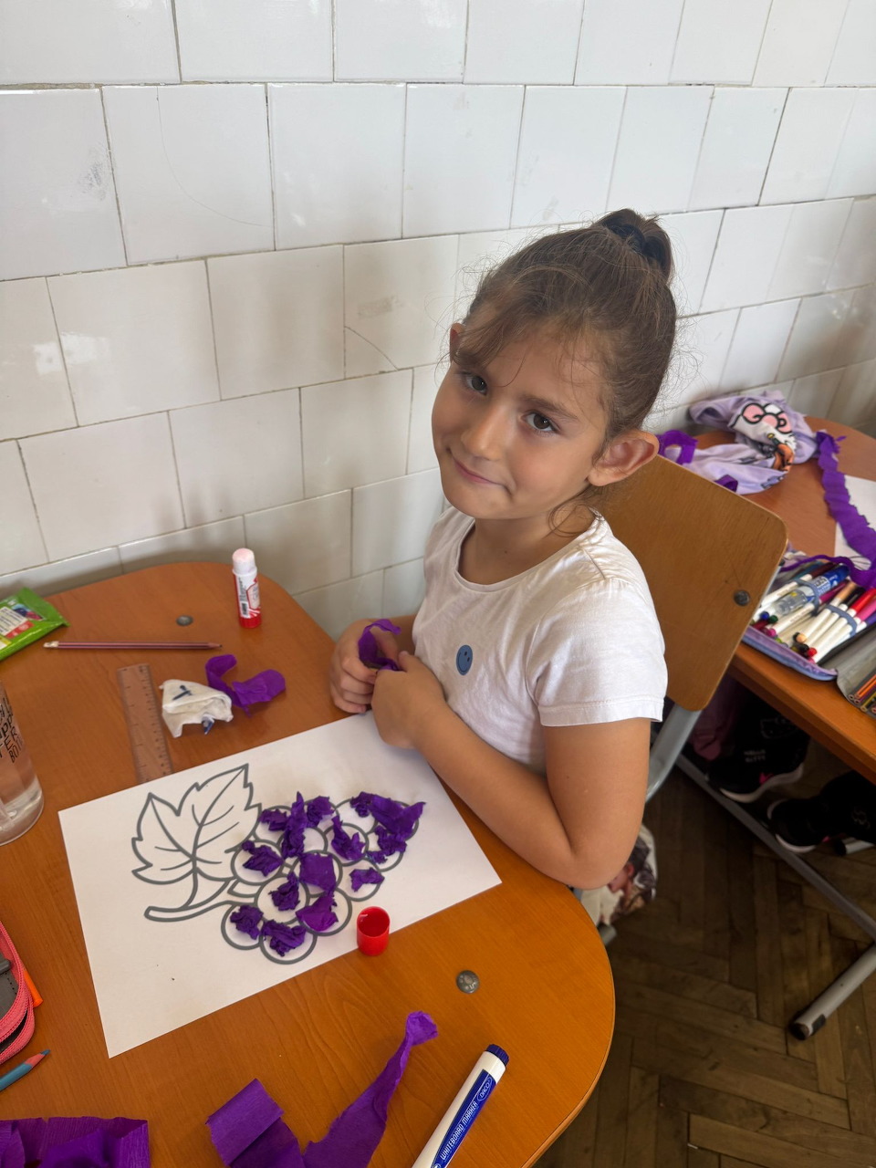 Young girl at a desk painting purple flowers on paper with art supplies nearby