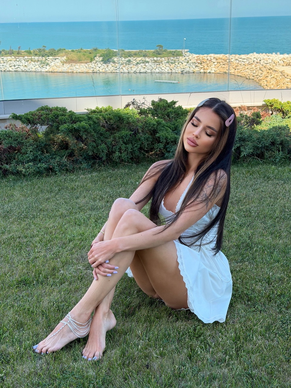 Woman with long dark hair sitting on green grass with ocean and greenery in the background