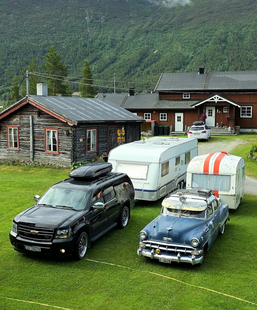 Black SUV and classic car with caravans parked on green lawn beside a log cabin in a Norwegian mountain valley.