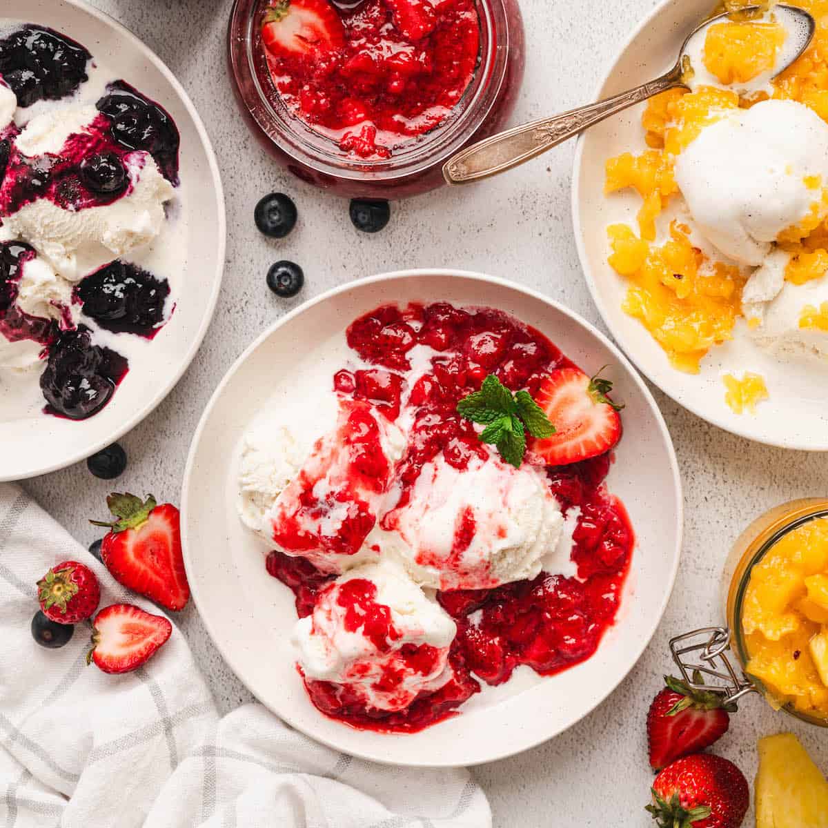 Three plates of ice cream with colorful fruit toppings including berries and mango on a white marble surface