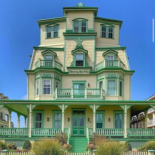 Pale yellow Victorian house with green trim and wraparound porch under a blue sky