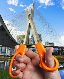 Hand holding orange scissors aligned with a cable-stayed bridge in a city scene