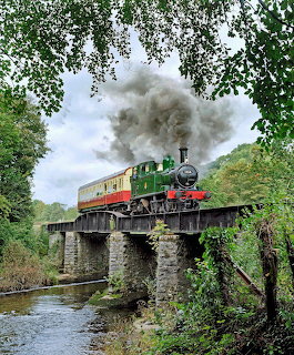 Steam train crossing stone bridge over river with trees and smoke plume