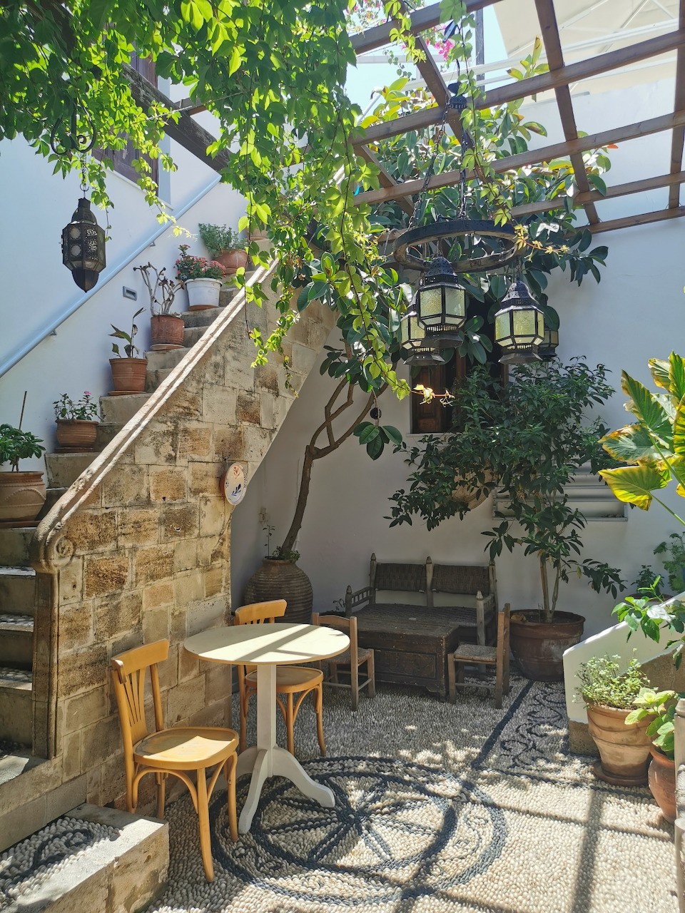 Stone courtyard with café table, chairs, hanging lanterns, potted plants and pergola in Lindos, Rhodes