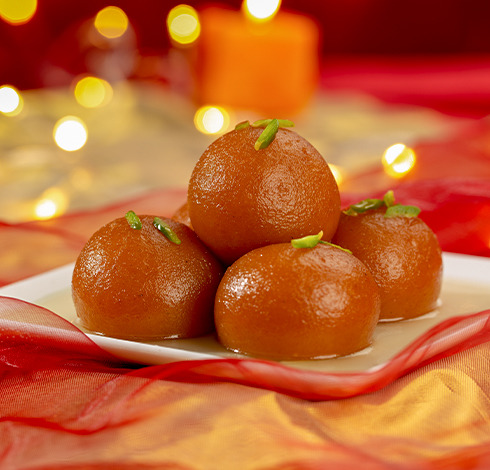 Five golden gulab jamun sweets garnished with pistachio on a white plate with warm bokeh background