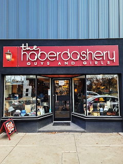 Haberdashery storefront with red sign, display windows, and a central door on a sidewalk