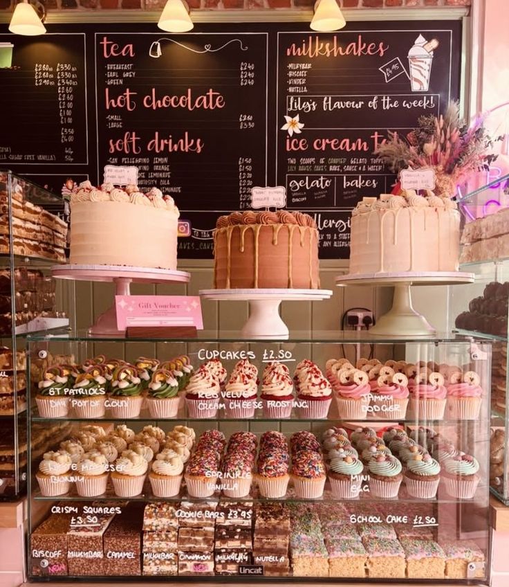 Bakery display case with three decorated layer cakes on top and rows of assorted cupcakes on lower shelves