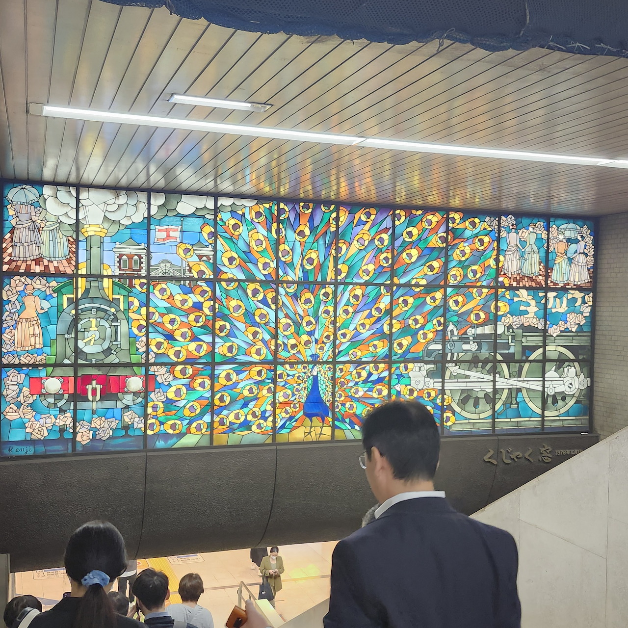 Colorful stained glass peacock mural in a subway station with people walking below