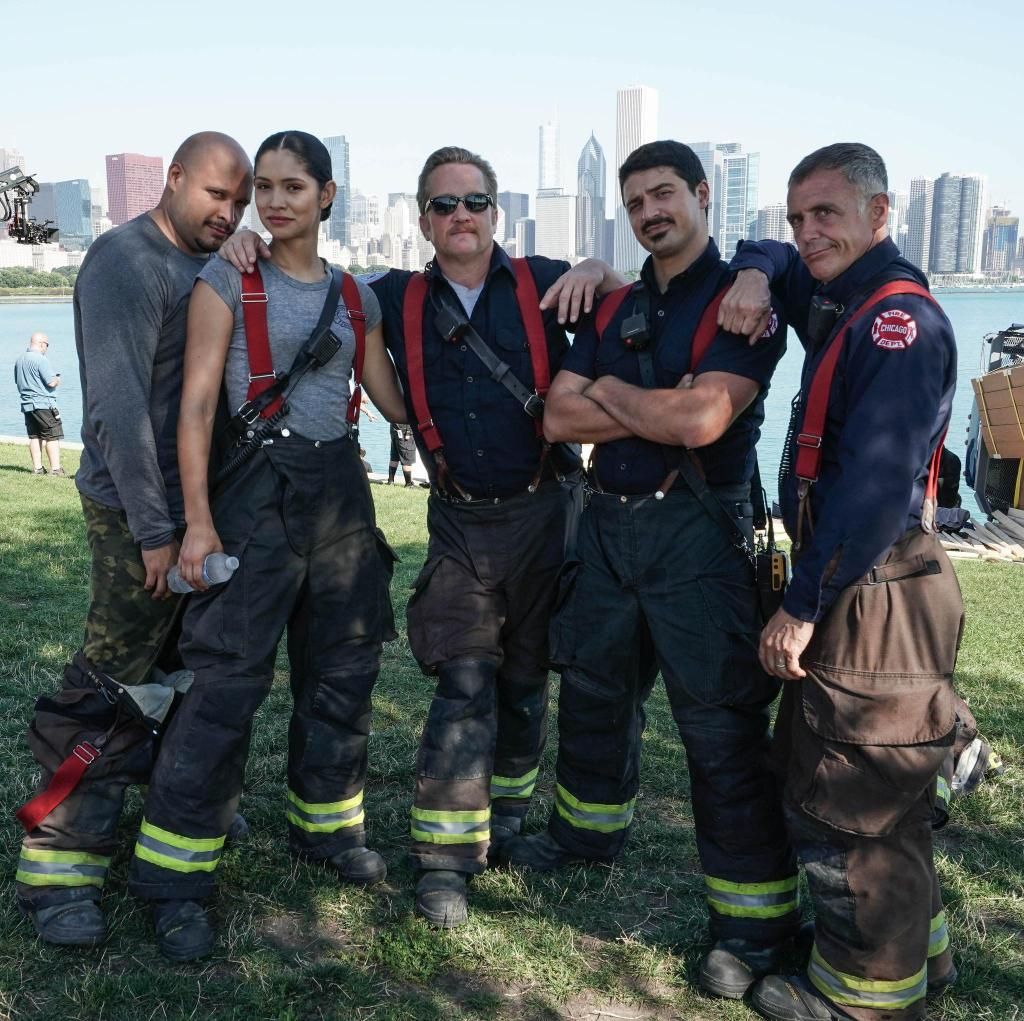 Five firefighters in gear posing on grass with a city skyline behind them