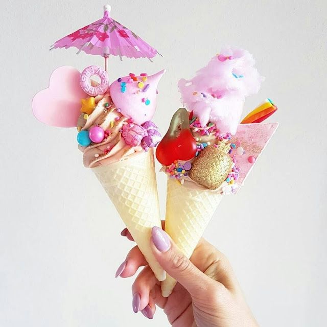 Hand holding two pastel ice cream cones topped with candy, sprinkles, and a pink umbrella