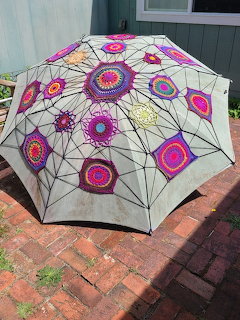 White umbrella adorned with vibrant crocheted floral motifs in pink, purple, orange and blue on brick patio