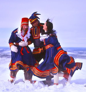 Three people in traditional blue, red, and yellow Sámi garments dancing on snow