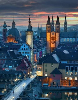 Twilight aerial view of Würzburg, Germany, with glowing church towers and historic rooftops reflected near a river.