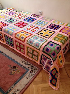 Colorful granny square crochet blanket covering a bed with a floral rug on the floor beside it