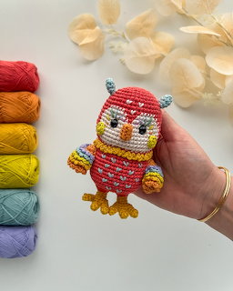 Hand holding a small beaded red owl with yellow feet and beak, yarn and flowers nearby