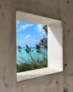 White window frame showing turquoise ocean view with coastal grasses and blue sky