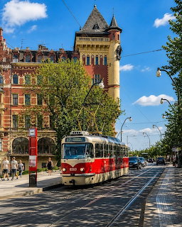 Red and white tram on Prague street with historic tower building and trees under blue sky
