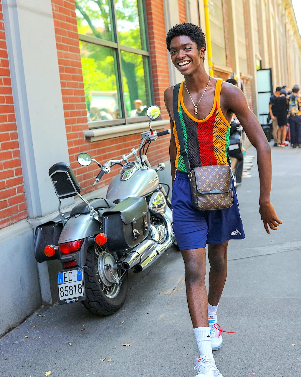 Man in rainbow striped vest and shorts standing beside motorcycle on urban street