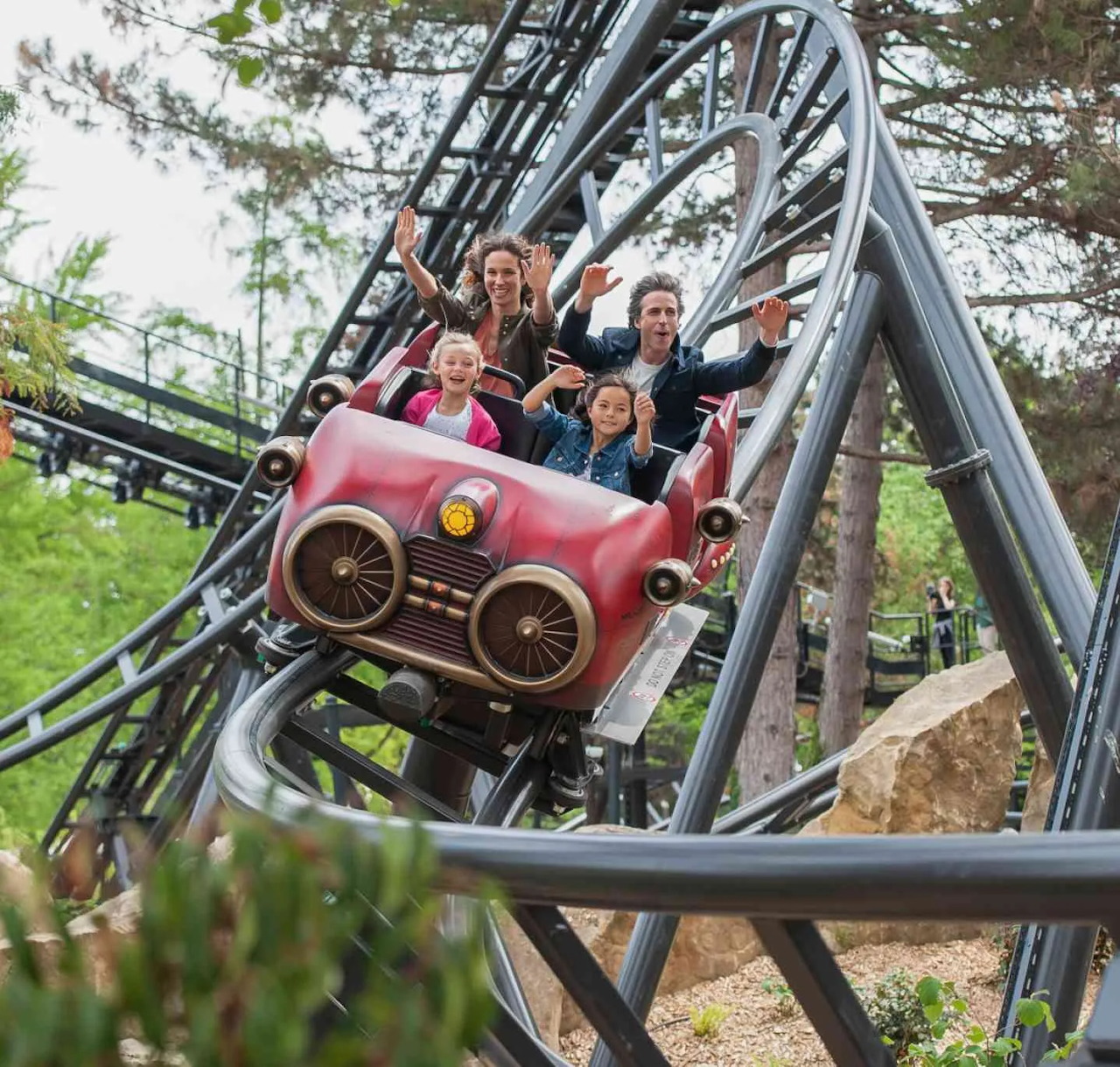 Family of four riding pink roller coaster car on elevated silver track in outdoor park setting