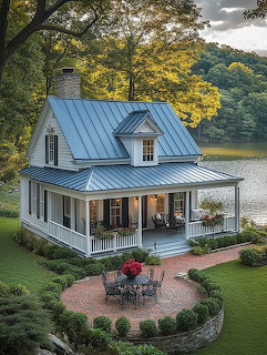 White cottage with blue roof and wraparound porch beside a lake surrounded by trees