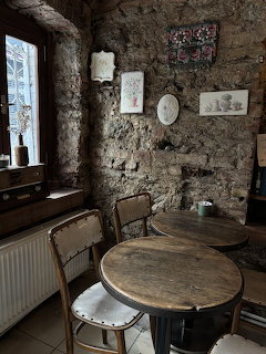 Rustic cafe interior with stone walls, wooden tables, chairs, and a window with lace curtains