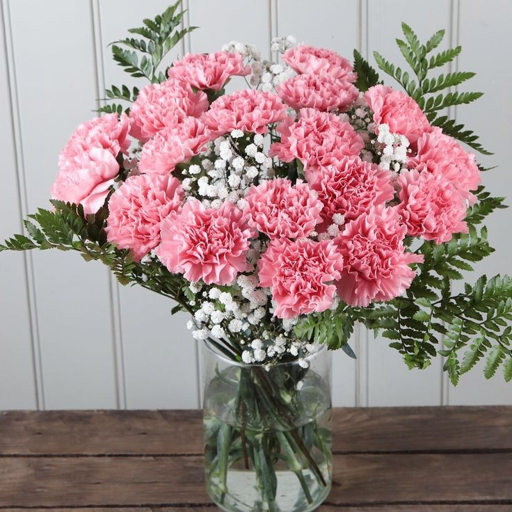 Pink carnations with baby's breath and fern leaves arranged in a glass vase on a wooden table