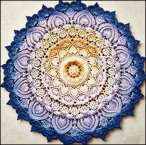 Circular crocheted doily with blue outer edge, violet and cream middle rings, and yellow center in mandala pattern