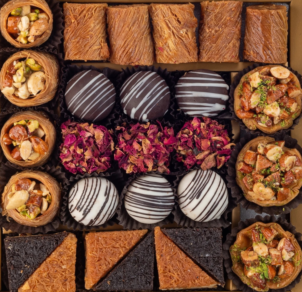 Assorted baklava and Middle Eastern sweets arranged in rows with chocolate drizzles and rose petals