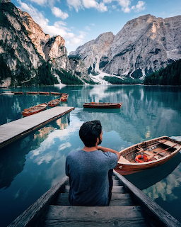 Person sitting on steps by a turquoise mountain lake with rowboats, dock, trees, and rocky peaks