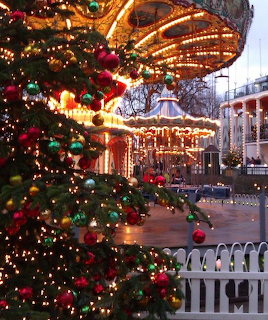 Large decorated Christmas tree with ornaments and lights next to an illuminated carousel in a city square at dusk