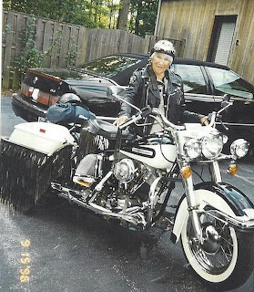 Elderly woman wearing leather jacket and helmet sitting on a parked cruiser motorcycle in a driveway