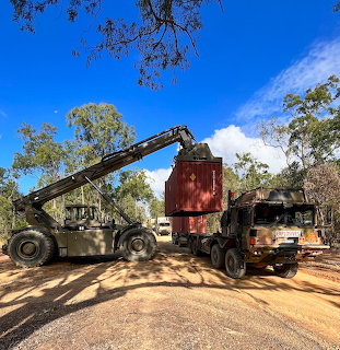 Loader lifting a red container onto a truck in a dirt yard with trees and blue sky