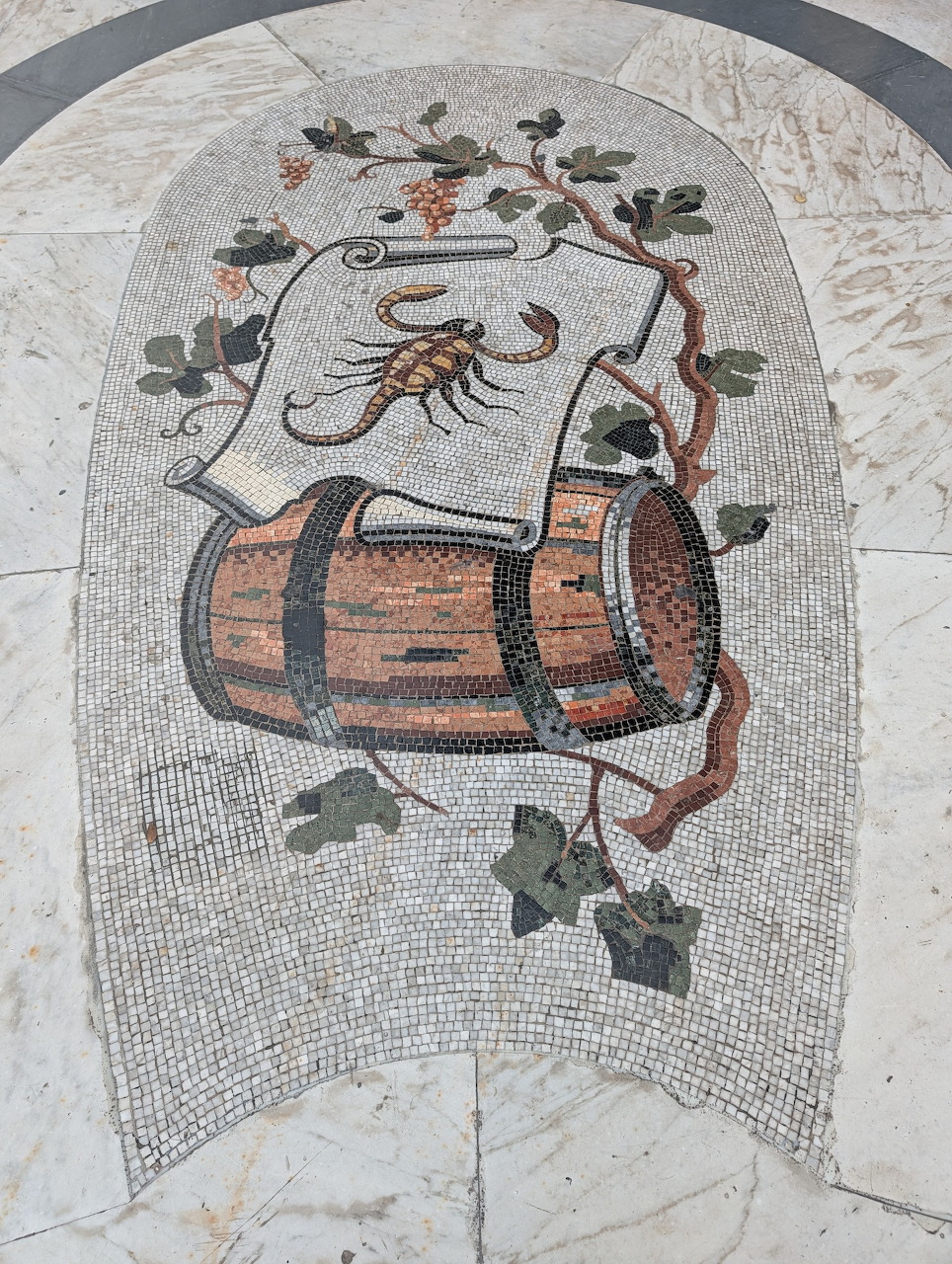 Floor mosaic of a scorpion, wooden barrel, and grapevine leaves framed by a stone arch