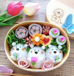 Cute bento box with rice ball faces, sushi rolls, vegetables, and flower-shaped food on a wooden table
