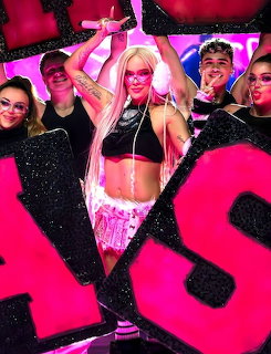 Female pop performer on stage with backup dancers, pink lighting, and large glittery letters