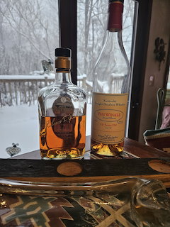 Two bourbon whiskey bottles on a wooden surface with glasses and a snowy landscape visible through a window