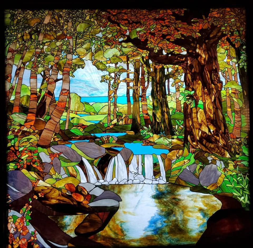 Stained glass artwork of a forest scene with waterfall, trees, rocks, and a reflective stream