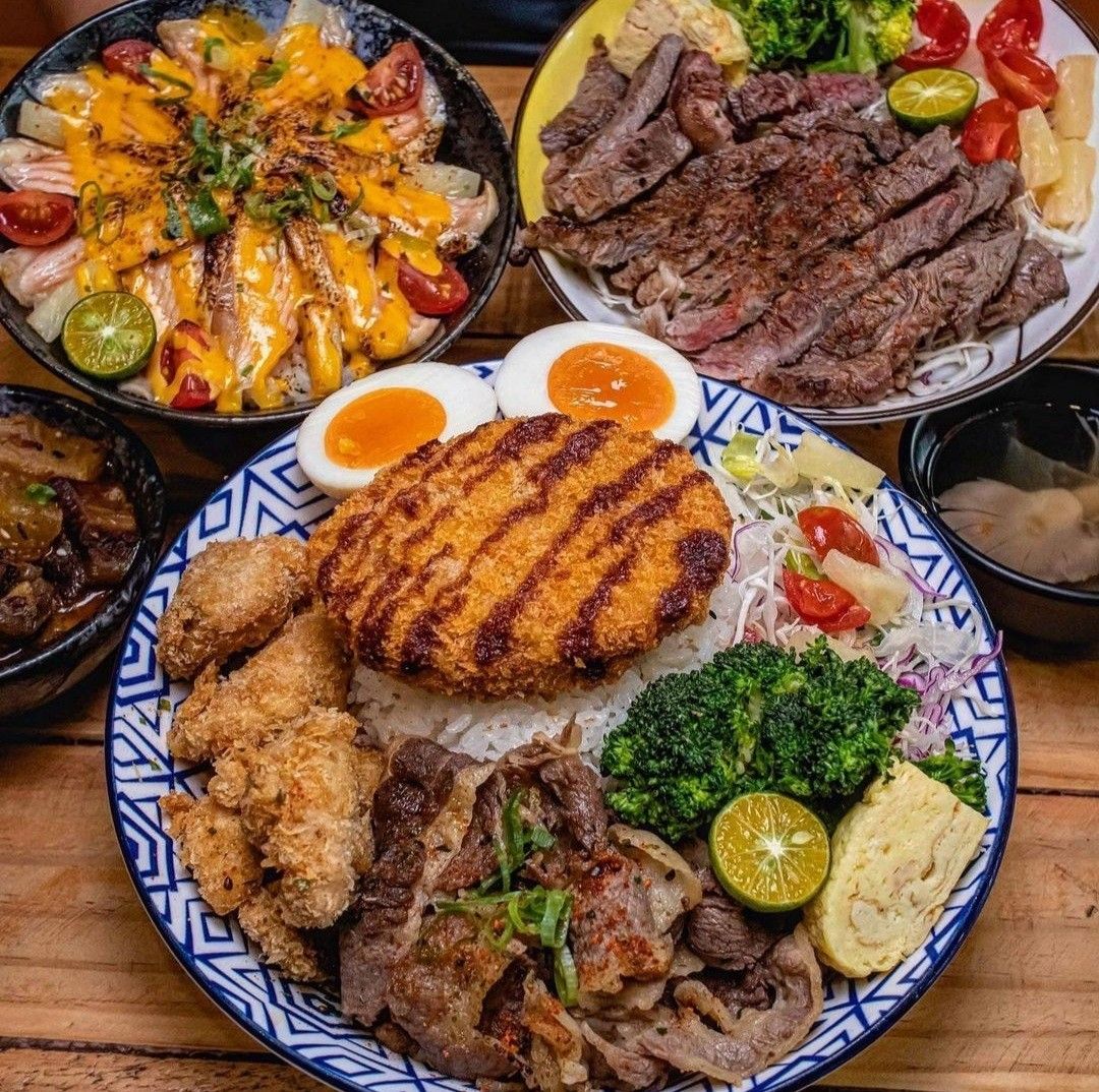 Multiple plates of grilled meats, breaded cutlets, rice, and colorful vegetables arranged on a wooden table