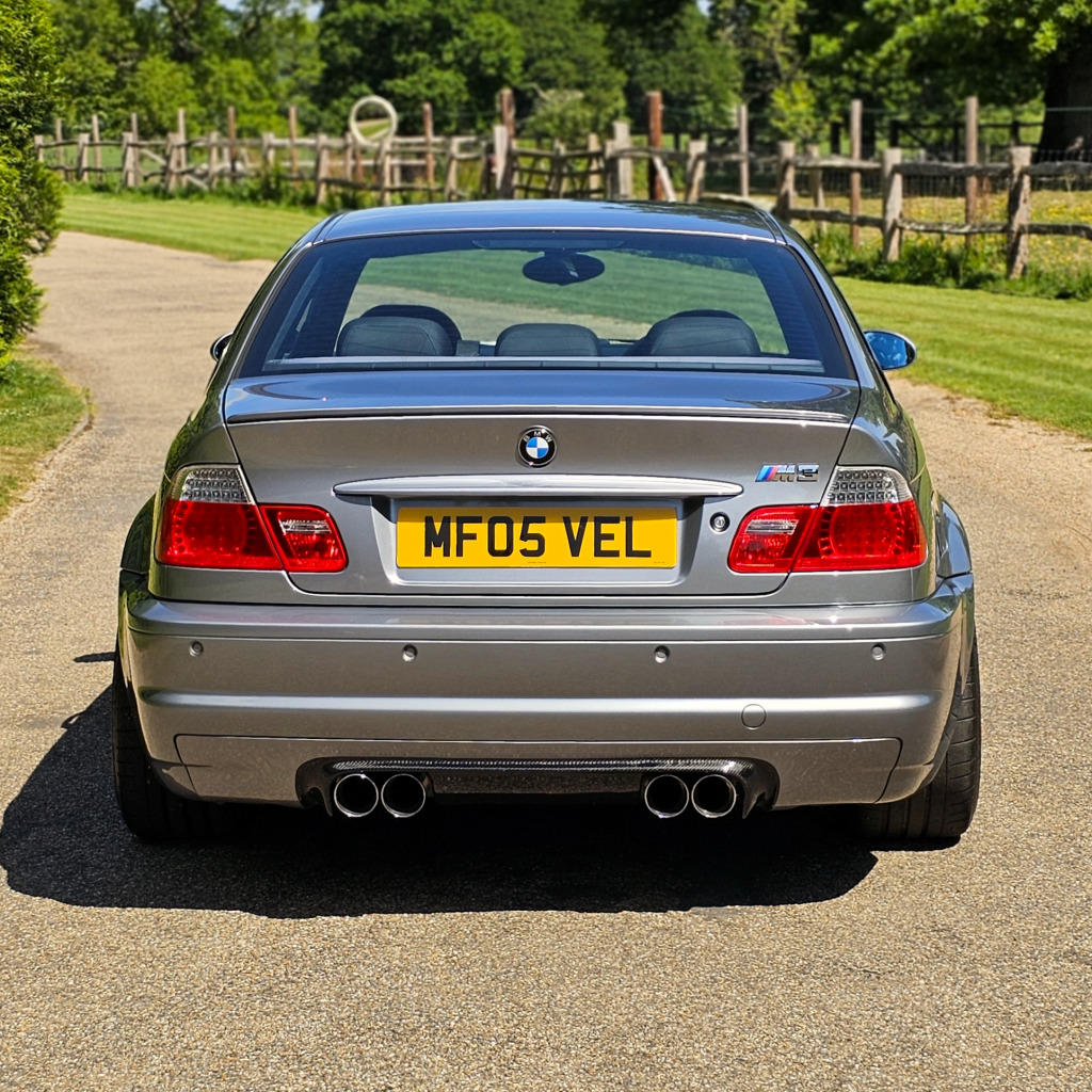 Rear view of a silver BMW car on a paved road with grass and a wooden fence behind it.
