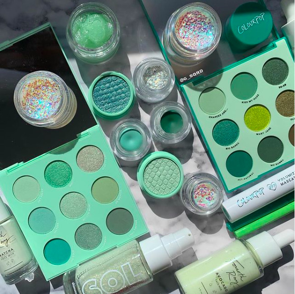 Green makeup flat lay with eyeshadow palettes, glitter jars, and cosmetic bottles