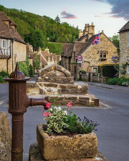 Historic Cotswolds village street with stone buildings, fountain and flowers at sunset