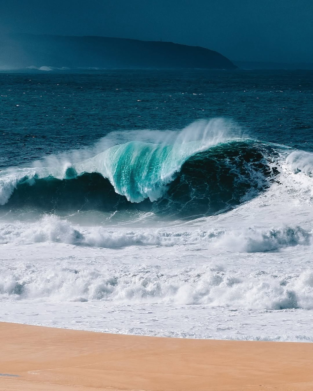 Large turquoise wave crashing near a sandy beach with a dark headland in the background