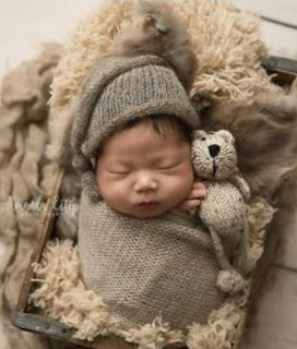 Sleeping newborn baby in gray knit wrap and hat holding a small knitted teddy bear in a wooden crate