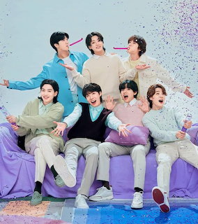 Seven BTS members posing together on a purple couch with confetti in pastel outfits