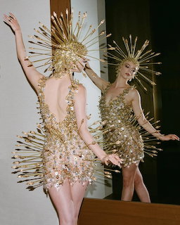 Woman in gold spiked couture dress and sunburst headpiece posing next to a mirror reflection