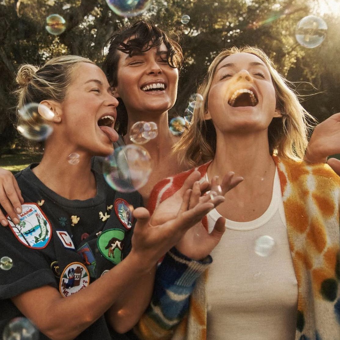 Three happy women laughing together outdoors with soap bubbles floating around them in sunlight