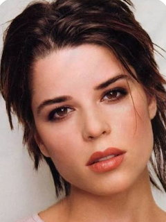 Close-up portrait of Neve Campbell with dark hair and soft makeup against a light background.