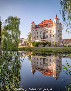 White castle with red roof and towers reflected in a pond surrounded by greenery and willow trees
