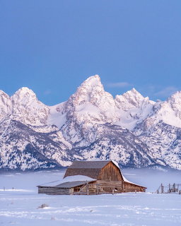 Wooden barn in snowy field with Grand Teton mountain peaks in background under blue sky