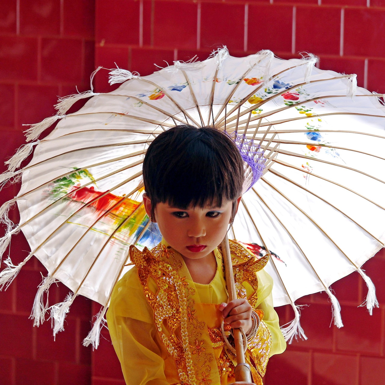Boy in yellow traditional dress holding white decorated umbrella against red wall